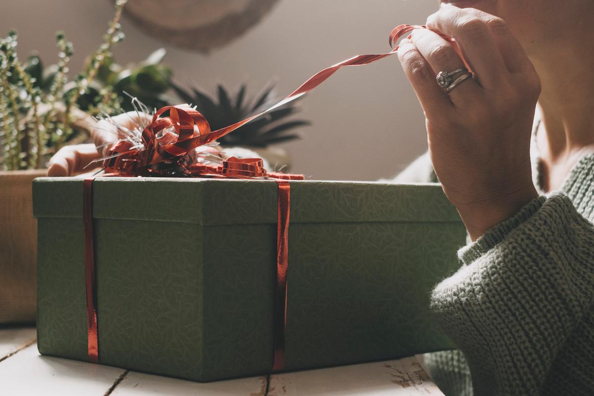  The image shows a person unwrapping a green gift box adorned with a shiny red ribbon and bow. The person’s hand is gently pulling the ribbon, and they are wearing a knitted sweater and a ring on their finger. In the background, plants can be seen, creating a cozy and natural ambiance.